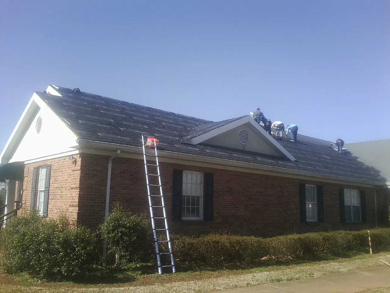 Crew tearing shingles off a brick ranch roof with a ladder set — Metal Roof Repair work by our crew in Waleska