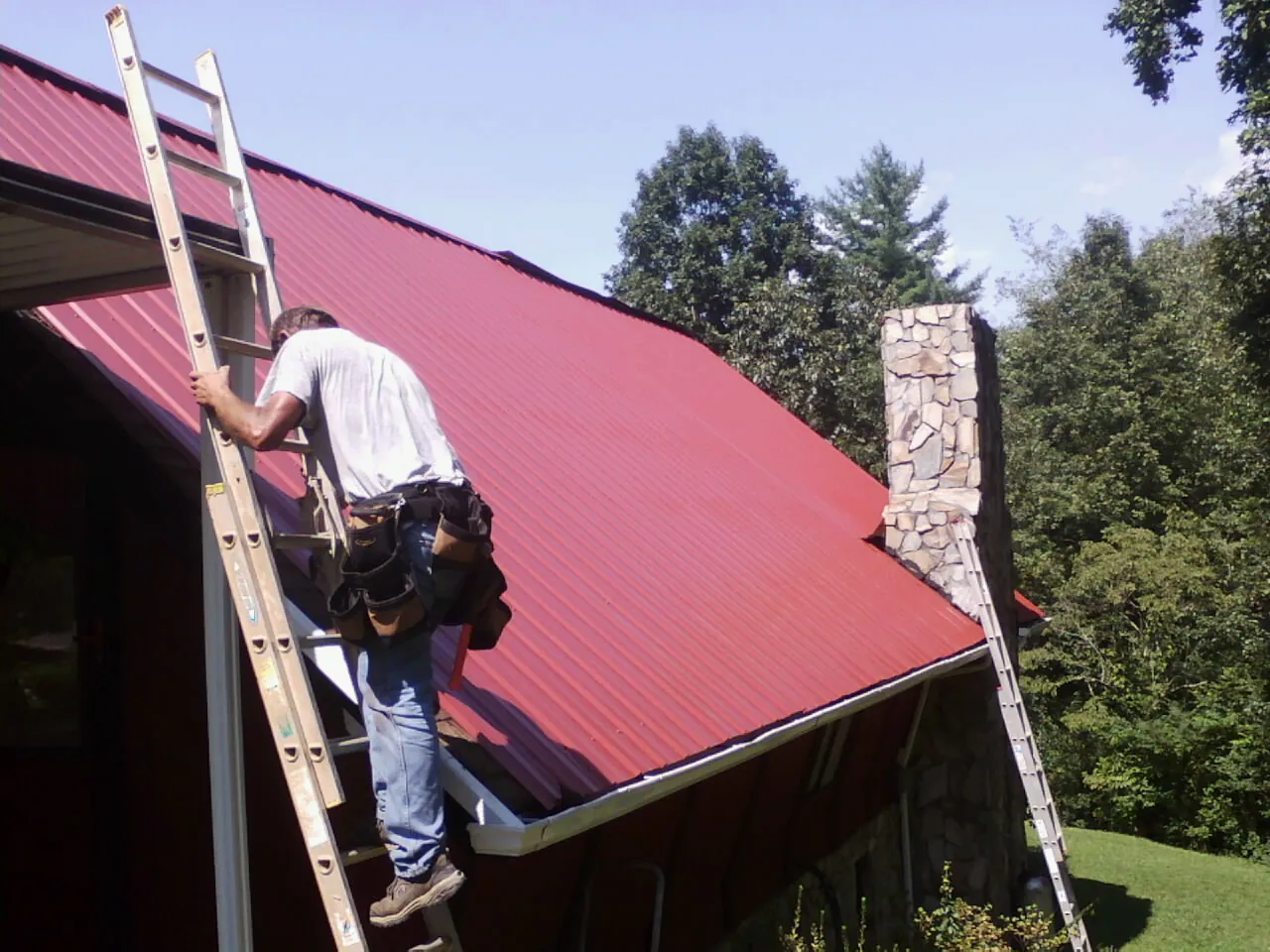 Roofer on a ladder beside a red metal roof and stone chimney — Soffit & Fascia Repair by Roofing Brothers in Waleska